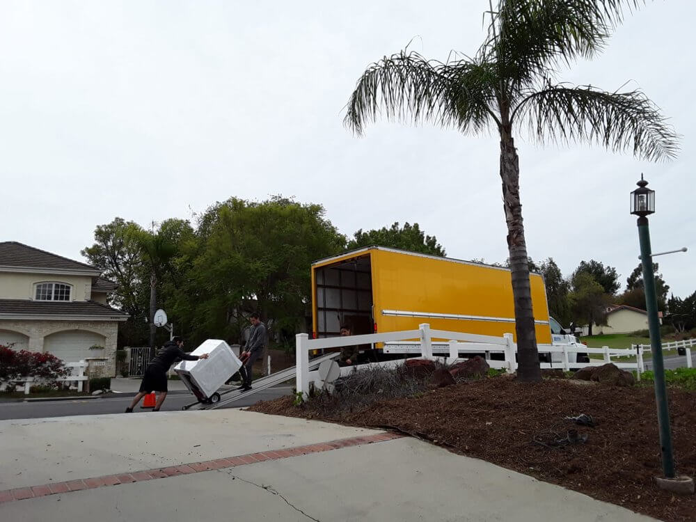 Movers loading a treadmill onto a yellow moving truck outside a suburban Los Angeles home